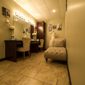 A photo of Alegria Gardens Reception Hall's well-lit dressing room with large mirrors, two chairs, and a bench.